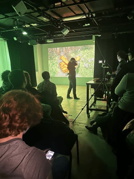 The presenter wearing monarch wings in front of the projected VR scene, audience in the foreground